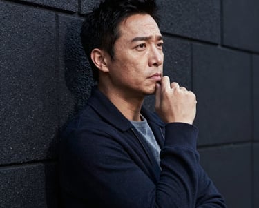 Candid shot of an actor in thought, leaning against a charcoal wall. High contrast lighting, North American urban setting.