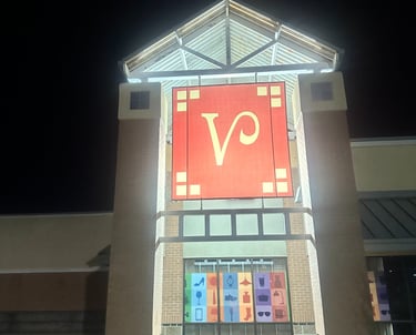 Illuminated shopping mall entrance at night featuring a large red logo sign and retail category icons.