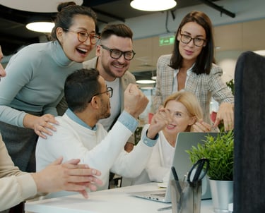 Diverse creative office team cheering and celebrating success while looking at a laptop screen together.