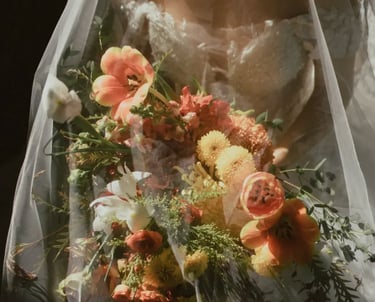 Novia con vestido de encaje bajo velo transparente sosteniendo un ramo en tonos naranja, blanco y verde.