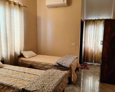 A bright PG room with two twin beds, tan curtains, and a wooden wardrobe on tiled floors.