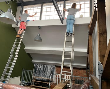 An in-progress living room, with a slanted ceiling and windows, and two painters working on ladders.