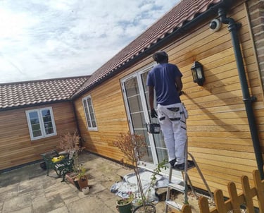 Exterior of wood-panelled bungalow, with a painter in-process of varnishing.