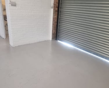 A corner of an industrial unit, with painted white brick walls, grey floor, and metal shutter door.