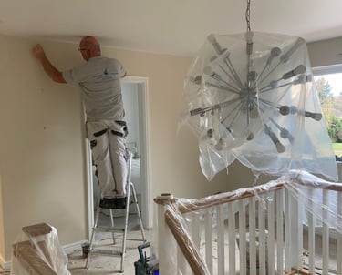 An in-process white landing/hallway, with sheet covered furniture and a painter on a ladder.