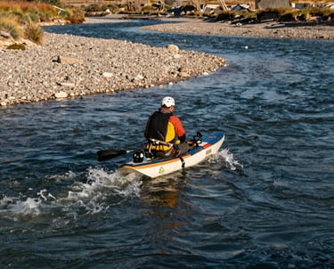 An outdoor action shot of a guide on a Montana river. The water is a deep slate blue, the banks are earthy off-white gravel, and the lighting is the bright, clear gold of a Montana morning.