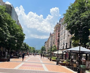 Sunny view of Vitosha Boulevard in Sofia, Bulgaria, featuring outdoor cafes, lush trees, and a mountain backdrop.