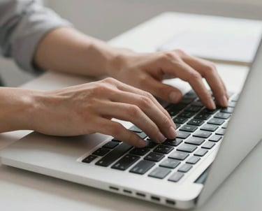 A focused shot of a person's hands typing on a laptop with a view of a clean, bright workspace behind them.