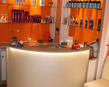 A modern hair salon reception desk with orange walls and glass shelves displaying professional beauty products.