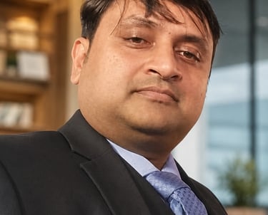 Professional portrait of a businessman wearing a black suit and blue tie in a corporate office setting.