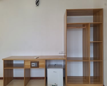 Modern hotel room furniture featuring a wooden desk with a digital safe, mini fridge, and open wardrobe shelving.