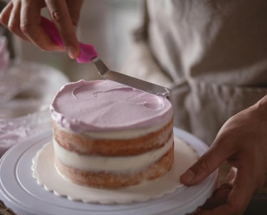 Baker using an offset spatula to spread purple frosting on a small layered sponge cake.