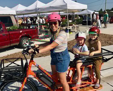 Woman stopped on a bike, posing with two young children seated in the back
