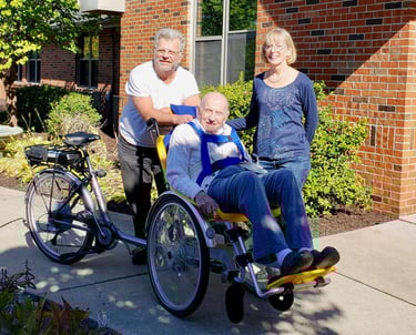 One person sitting in the Giving Rides bike with two people standing behind them smiling