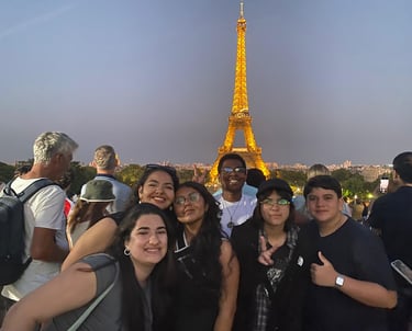 A imagem mostra um grupo de estudantes em frente à Torre Eiffel iluminada, em Paris.