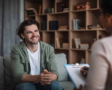 A smiling man sits on a sofa during a therapy session with a mental health professional.
