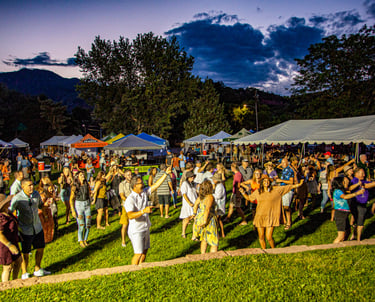 Crowd dancing at an outdoor summer music festival with event tents and mountains at sunset.