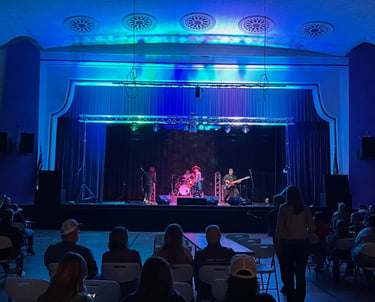 Live band performing on stage at an Memorial Hall with blue and green stage lighting and audience.