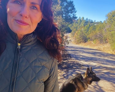 A woman in a quilted jacket hiking with her German Shepherd on a sunny trail.