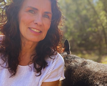 A smiling woman with curly brown hair wearing a white top outdoors with her dog.