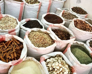 Large white sacks filled with various bulk spices, dried herbs, and roots in an open-air market stall.