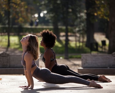 Two women practice upward facing dog yoga pose outdoors in a sunny park.
