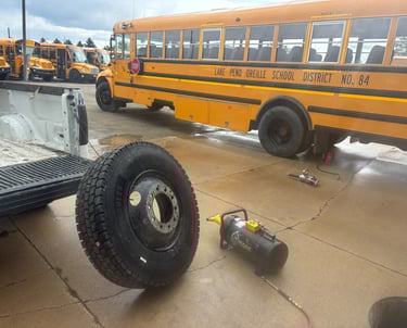 A mechanic services a yellow school bus tire and wheel on a wet pavement lot.