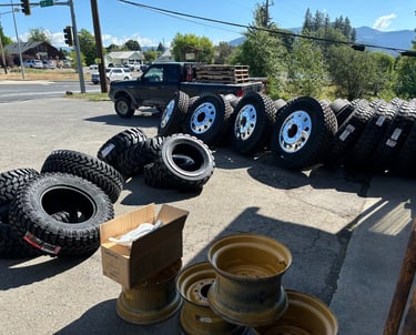 A collection of heavy-duty truck tires and chrome rims displayed on asphalt at a tire service shop.