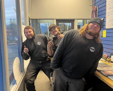 Three cheerful auto shop mechanics in black hoodies posing and giving thumbs up in a garage office.