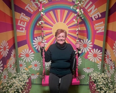 Photo of Debbie, a white woman with short red & blonde hair, on a flowery swing in front of a sign: "Let the Sunshine In."