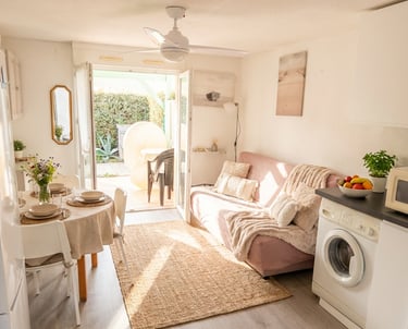 Bright studio apartment with a cozy pink sofa, dining table, and kitchen area leading to a sunny outdoor patio.