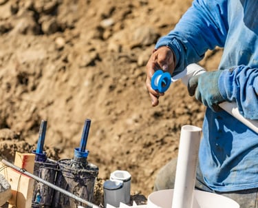 A plumber in a blue shirt is taping PVC piping on a construction site.