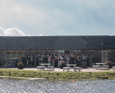 Wedding guests posing for a group photo outside a rustic stone barn venue by a scenic pond.