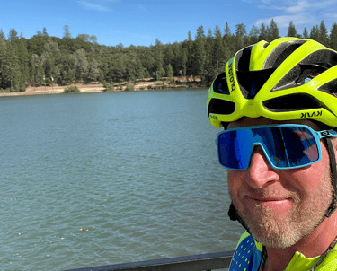 A picture of Justin in cycling gear standing next to a body of water (lake).