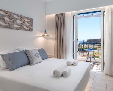 Modern Mediterranean hotel room with white bed linens, blue pillows, and a balcony overlooking the sea.