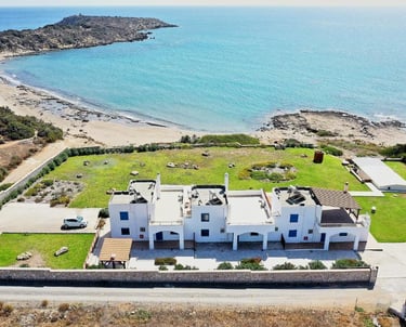 Aerial view of a white beachfront villa with blue shutters on a scenic Mediterranean coastline.