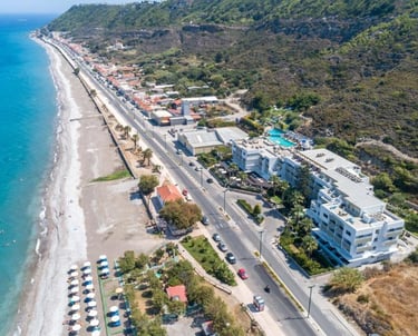 Aerial view of a coastal resort and sandy beach in Rhodes, Greece, with turquoise sea and green hills.