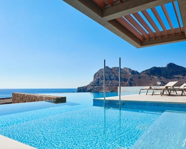 Luxury hotel infinity pool with ocean views, mountains, and lounge chairs on a sunny terrace.