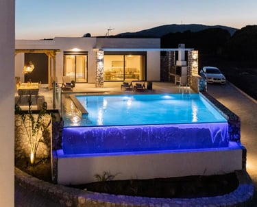 Luxury modern villa at dusk featuring an illuminated infinity pool with blue lighting and outdoor dining area.