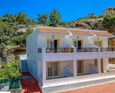 Two-story white Mediterranean villa with balconies nestled against a rocky hillside under a clear blue sky.