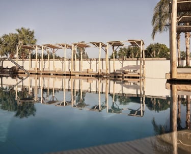 Luxury resort swimming pool with wooden cabanas and palm tree reflections on calm water.