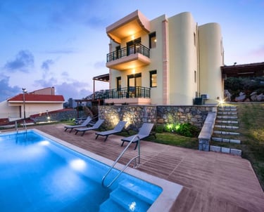 Luxury three-story villa with a lit swimming pool, sun loungers, and scenic views at dusk.