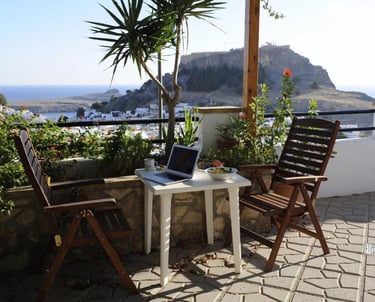 Laptop on a terrace table with wooden chairs overlooking Lindos Acropolis and the sea.