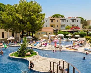Families relaxing by a large outdoor resort swimming pool with sun loungers and palm trees.