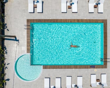 Aerial view of a woman floating in a luxury resort swimming pool with a sun deck and white lounge chairs.