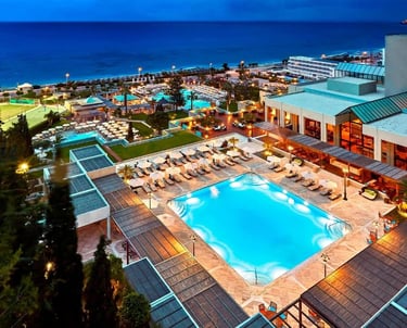 Luxury beach resort with illuminated swimming pools and tennis courts at dusk overlooking the ocean.