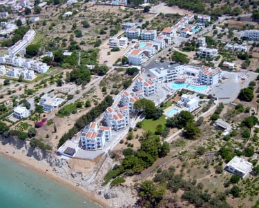 Aerial view of a coastal Greek island resort with white villas and swimming pools near the beach.