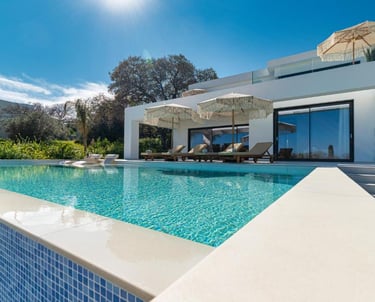 Modern luxury villa with a private infinity pool, sun loungers, and umbrellas under a clear blue sky.