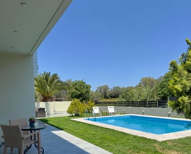 Luxury villa backyard featuring a private swimming pool, green lawn, and patio seating under a clear blue sky.