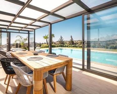 Modern covered patio with wooden dining table and glass sliding doors overlooking a luxury swimming pool.